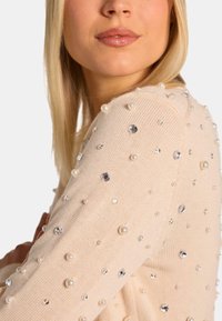 Beige Strickpullover mit runden Perlen und Juwelen, die die Ärmel akzentuieren. Weicher Stoff, taillierte Passform und Rundhalsausschnitt.