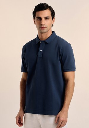 Polo de color azul marino hecho de algodón texturizado, con un cuello de tipo polo, tapeta de dos botones y mangas cortas. Detalle del logo sutil en el pecho.