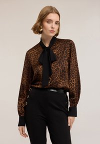 Bluzka w panterkę z brązowego i czarnego jedwabiu z wiązanym kołnierzykiem, w połączeniu z czarnymi spodniami z wysokim stanem ozdobionymi złotymi guzikami.
