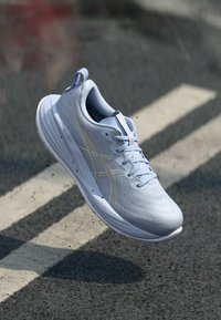 Sneaker de sport bleu clair avec un matériau texturé, un col rembourré, des lacets, des accents argentés et une semelle rembourrée, flottant au-dessus d'une surface.