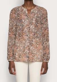 Blouse à manches longues à motifs floraux dans des tons de rose, brun et beige, portée avec un pantalon écru, présentant un col en V et une fermeture à boutons.