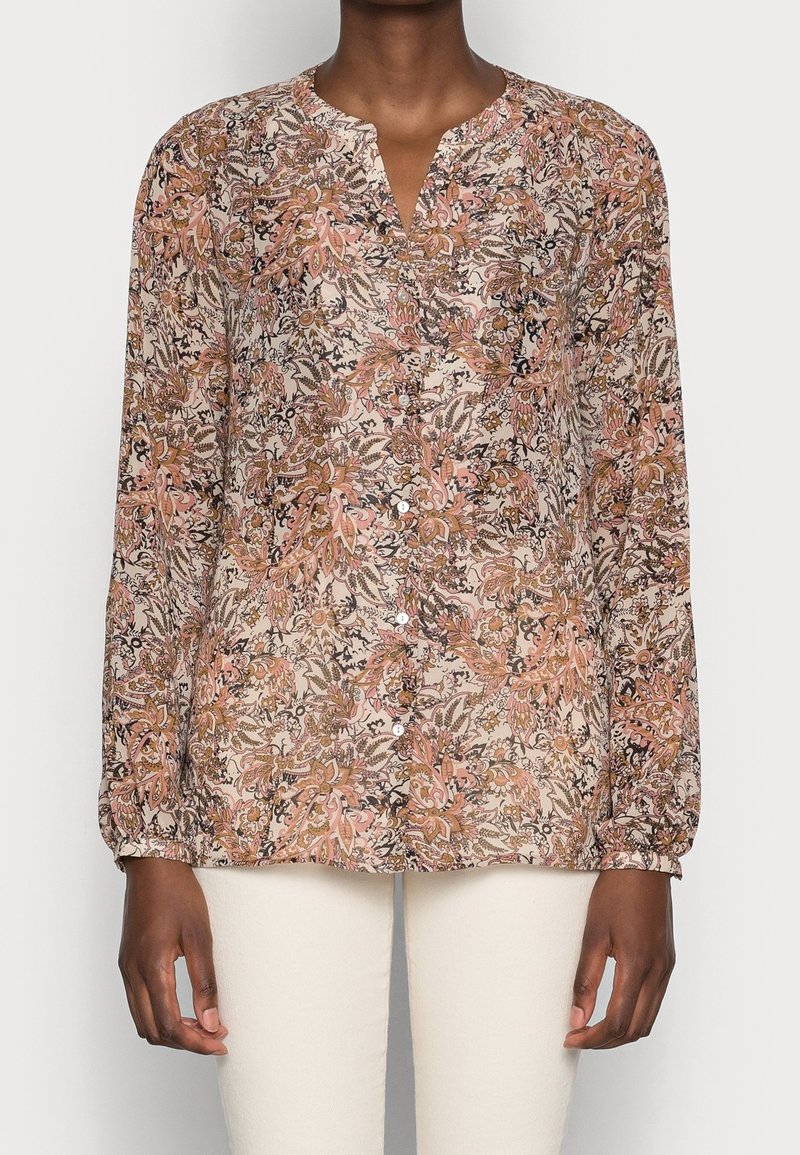 Blouse à manches longues à motifs floraux dans des tons de rose, brun et beige, portée avec un pantalon écru, présentant un col en V et une fermeture à boutons.
