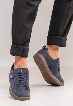 Osoba nosząca ciemnoniebieskie zamszowe trampki z brązowymi podeszwami i podwiniętymi czarnymi spodniami, stojąca na jasnoszarej podłodze.