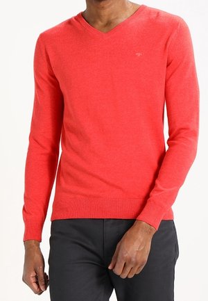 Man wearing a fitted red V-neck sweater with long sleeves and black pants, standing against a white background.