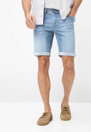 Man draagt lichtblauwe opgerolde denim shorts, wit overhemd en beige suède loafers, staand tegen een effen achtergrond.