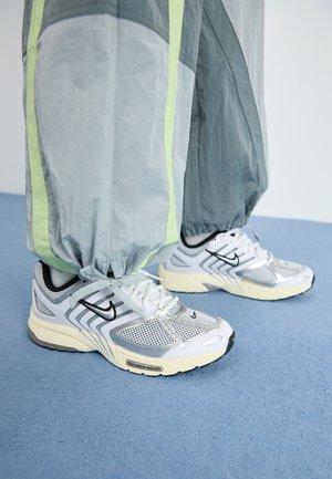 Graue und limettengrüne Trainingshose mit gerafften Bündchen, getragen über weißen und silbernen Sneakers mit schwarzen Akzenten und Netzdetails.