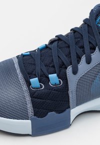 Gros plan d'une chaussure de sport bleu marine avec tissu matelassé, passants de lacets bleus et bout ainsi que panneau latéral gris texturé.