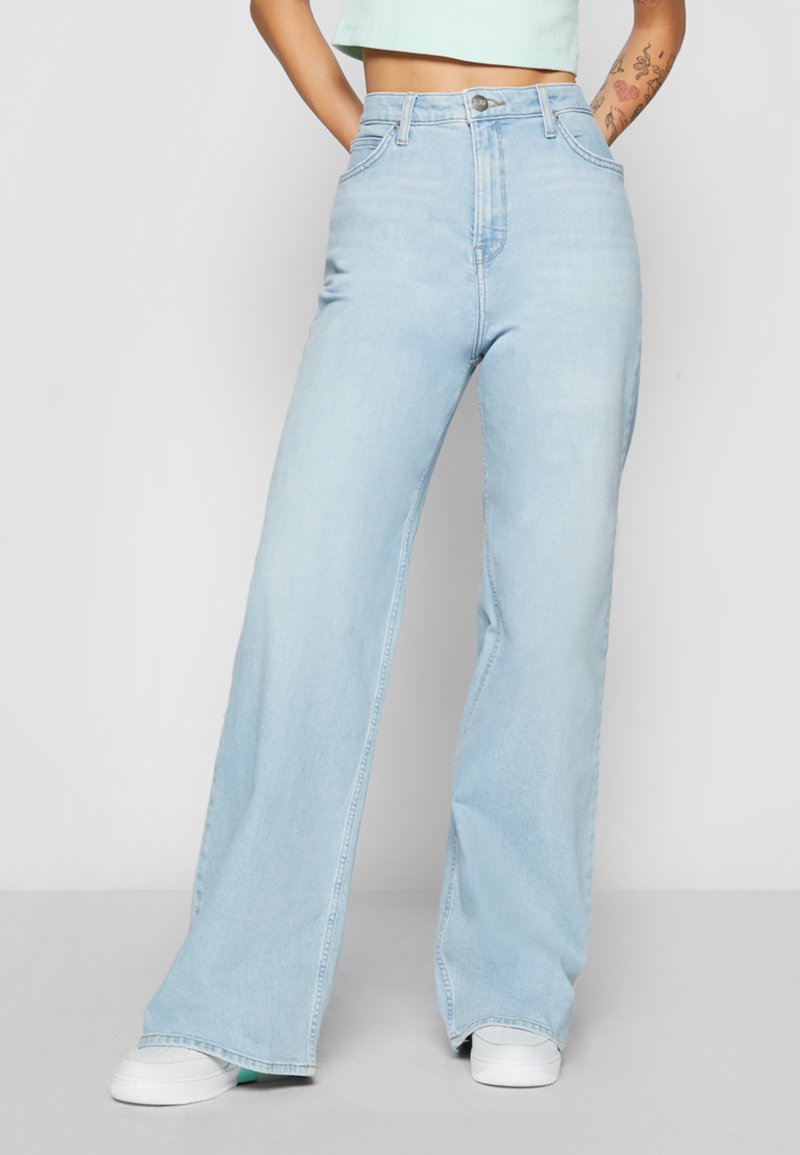 Personne portant un jean taille haute bleu clair à jambes larges, les mains dans les poches et des baskets blanches, montrée de la taille jusqu'aux pieds.