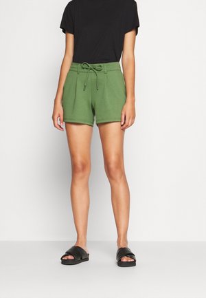 Persona vistiendo pantalones cortos de cordón de color verde oliva, camiseta negra y sandalias negras tipo slide, de pie frente a un fondo blanco liso.