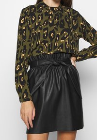 Blusa a maniche lunghe verde oliva con stampa leopardata nera e beige; abbinata a una gonna nera in ecopelle con dettaglio della cintura annodata.
