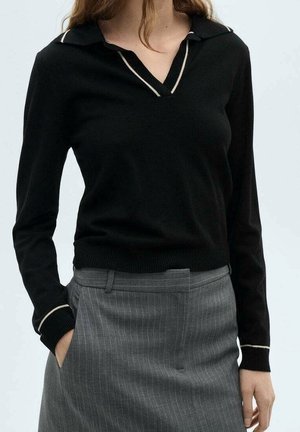 Femme portant un pull noir à manches longues avec un liseré blanc au col et aux poignets, associé à un pantalon gris à rayures fines taille haute, la main dans la poche.
