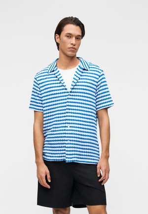 Junger Mann, der ein blau-weiß gestreiftes Kurzarmhemd über einem weißen T-Shirt und schwarzen Shorts trägt, vor einem einfarbigen Hintergrund stehend.