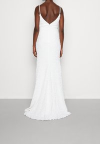 Vit kvällsklänning med en figursydd silhuett, låg V-rygg och tunna axelband. Strukturerad tyg med skimrande accenter, golvlång design.