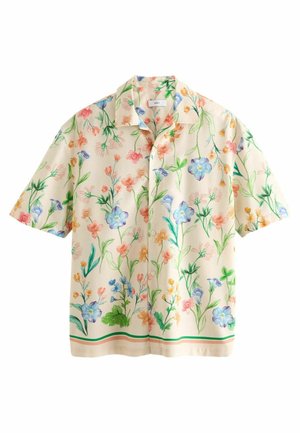 Homme portant une chemise crème à manches courtes avec un imprimé floral pastel, un pantalon bleu clair, des sandales noires, debout à l'extérieur devant une fenêtre et un mur de briques.