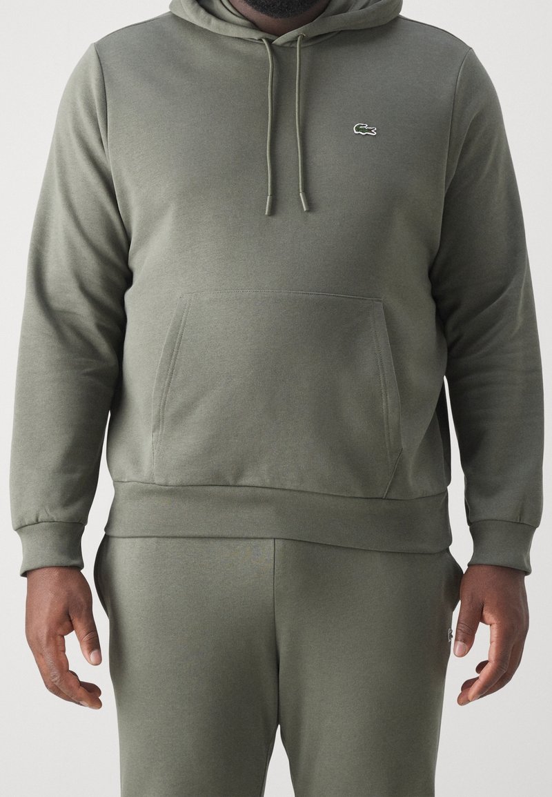 Homme portant un sweat à capuche vert olive et un pantalon de survêtement assorti, debout avec les bras détendus, présentant un petit logo crocodile sur la poitrine.