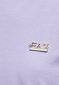 Καρφίτσα με το λογότυπο Gold EA7 Emporio Armani σε υφάσμα ανοιχτού μοβ χρώματος.