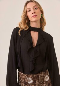 Blusa negra con volantes, cuello alto y detalle de apertura, hecha de tela texturizada. La modelo lleva puestos pantalones con estampado y se ven pendientes de aro verdes.