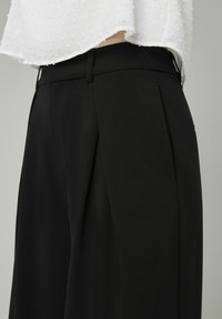 Pantalons noirs ajustés à taille haute, avec un devant plissé et des poches latérales. Le tissu a une texture lisse et structurée.