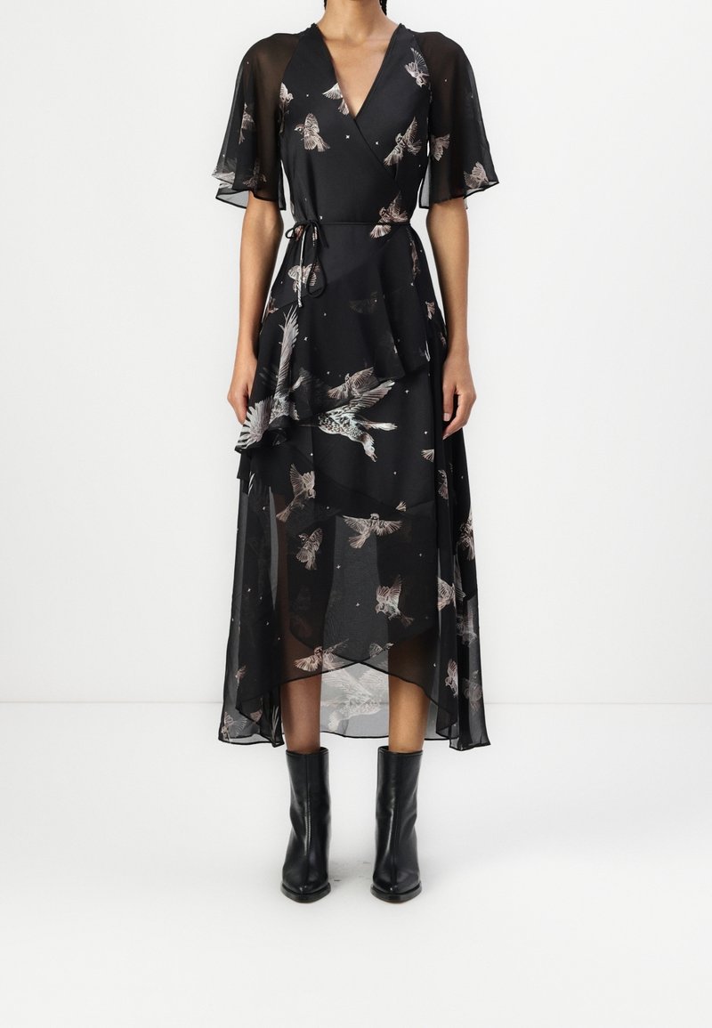 Femme portant une robe portefeuille noire transparente avec des imprimés d'oiseaux et des manches flottantes, associée à des bottines noires, debout sur un fond blanc.