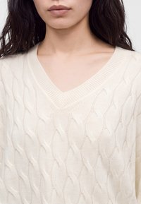 Femme aux longs cheveux foncés portant un pull crème à col en V en maille torsadée dévoilant le décolleté et le haut de la poitrine.