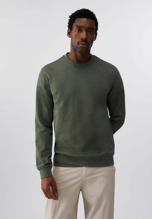 Mann trägt ein olivgrünes langärmeliges Sweatshirt und beige Hosen, steht vor einem schlichten hellgrauen Hintergrund.