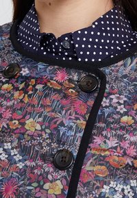 Takki, jossa on kukkakuosi ja mustat reunat sekä napit, yhdistettynä tummansiniisiin pilkkupaitaan. Sisältää eloisia värejä ja monimutkaisia yksityiskohtia.