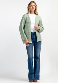 Jasnozielony dzianinowy kardigan o luźnym fasonie, z otwartym przodem i długimi rękawami, zestawiony z niebieskimi spodniami dzwonami i brązowymi botkami za kostkę.