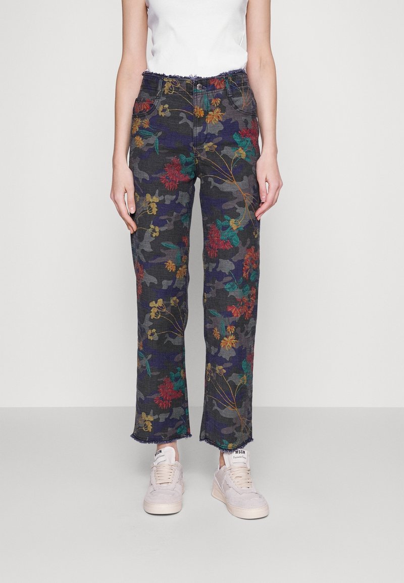 Femme portant un pantalon droit taille haute à imprimé camouflage floral foncé avec des ourlets effilochés, un haut sans manches blanc et des baskets blanc cassé.