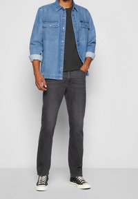 Blaues Jeanshemd mit zwei Brusttaschen, getragen über einem schwarzen T-Shirt, kombiniert mit dunkelgrauen Jeans und schwarzen High-Top-Sneakern.