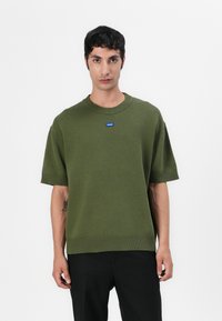 Olive green knitted sweater with short sleeves, ribbed neckline and hem, featuring a small blue logo label on the chest.