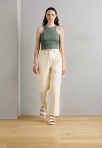 Un top a canottiera verde aderente abbinato a pantaloni beige a vita alta e sandali bianchi. L'outfit presenta un design semplice e moderno.