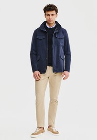 Granatowa kurtka z dwoma kieszeniami na piersiach i otwartym kołnierzem, zestawiona z ciemnym swetrem, beżowymi spodniami i jasnobrązowymi sneakersami.