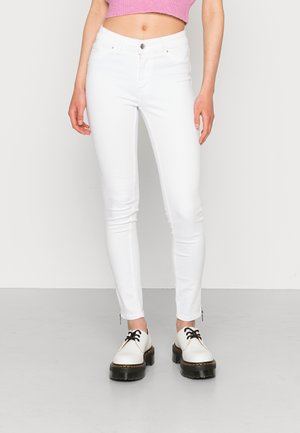 Jeans brancos justos com uma cintura alta e desbotamento subtil, apresentando detalhes em zíper nos tornozelos. Combinados com sapatos brancos e atacadores pretos.