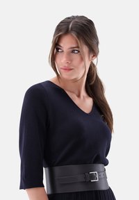 Marinefarbener V-Ausschnitt-Pullover aus weichem Stoff, kombiniert mit einem breiten schwarzen Ledergürtel, der eine silberne Schnalle und gestickte Details aufweist.