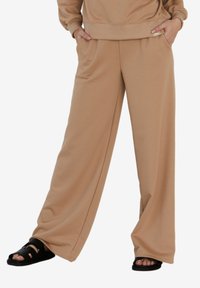 Beżowe spodnie typu wide-leg wykonane z miękkiego materiału z bocznymi kieszeniami; noszone z czarnymi sandałami. Prosty design i wygodny krój.