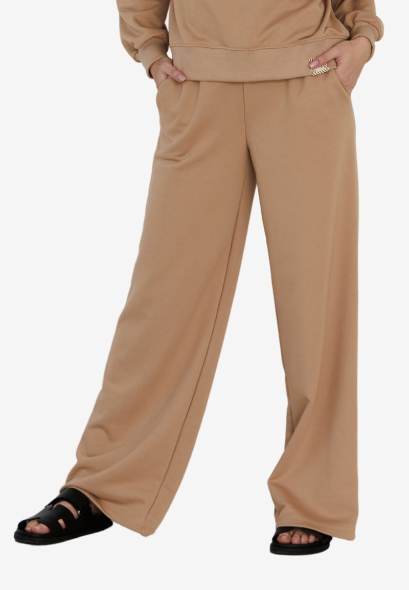 Beżowe spodnie typu wide-leg wykonane z miękkiego materiału z bocznymi kieszeniami; noszone z czarnymi sandałami. Prosty design i wygodny krój.