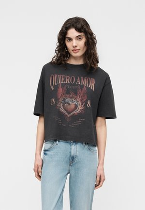 Femme aux cheveux bouclés et foncés portant un t-shirt noir à motif "Quiero Amor Tour 1981" et un jean bleu clair, debout devant un fond uni.