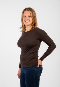 Top marrone a maniche lunghe realizzato in tessuto elasticizzato, design aderente, abbinato a jeans blu scuro, esposto su uno sfondo bianco uniforme.
