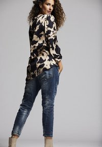 Langärmlige Bluse in Schwarz mit cremefarbenem abstraktem Muster, kombiniert mit abgewetzten blauen Jeans und beigefarbenen Ankle Boots. Lockige Haare sichtbar.