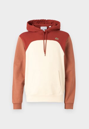 Capuchon sweatshirt met een crèmekleurig lichaam, rode accenten en bruine mouwen. Heeft een kangoeroe-zak en een klein groen logo op de borst.