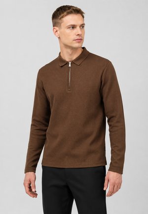 Jonge man die een bruine gestructureerde poloshirt met lange mouwen en korte ritssluiting aan de voorkant draagt, staand voor een effen lichte achtergrond.