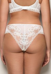 Kobieta w białym koronkowym biustonoszu i dopasowanych koronkowych majtkach w kwiaty, pokazana z tyłu na jednolitym tle.