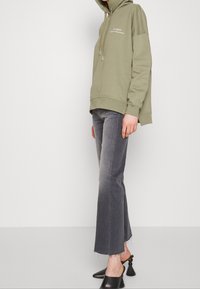 Sweat à capuche vert olive avec cordon de serrage, poche avant et texte imprimé, associé à un jean évasé gris et des mules noires ornées de détails à pampilles.