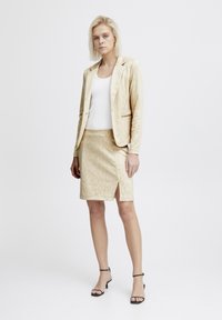 Ensemble de blazer et jupe beige avec un motif animal subtil, forme ajustée et fente latérale. Comprend un haut blanc et des escarpins noirs à brides.