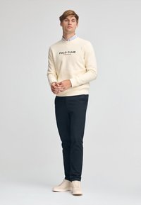 Sudadera color crema con gráfico "POLO CLUB", combinada con pantalones oscuros y zapatillas beige. El conjunto tiene un ajuste casual y estructurado.