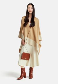 Pončo v barvě velbloudí s krémovým lemem, doplněné širokými bílými kalhotami, červenou kabelkou se vzorem krokodýlí kůže a odpovídajícími kozačkami po kolena.