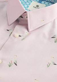 Chemise en tissu rose présentant un motif de hérons gris parmi des roseaux verts, avec un bouton blanc et un imprimé floral bleu à l'intérieur du col.