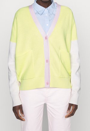 Personne portant un gilet jaune pastel avec des boutons roses et des manches blanches, par-dessus une chemise à col bleu clair et un pantalon rose pâle.
