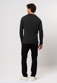 Cămașă neagră cu mânecă lungă, guler rotund, confecționată dintr-un material neted, asortată cu pantaloni negri și adidași albi, văzută din spate.