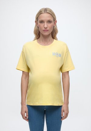 Nosečnica s svetlimi lasmi, oblečena v rumeno majico z napisom »MAMA« in cvetličnim vzorcem ter modrim jeansom, stoji pred enobarvnim ozadjem.